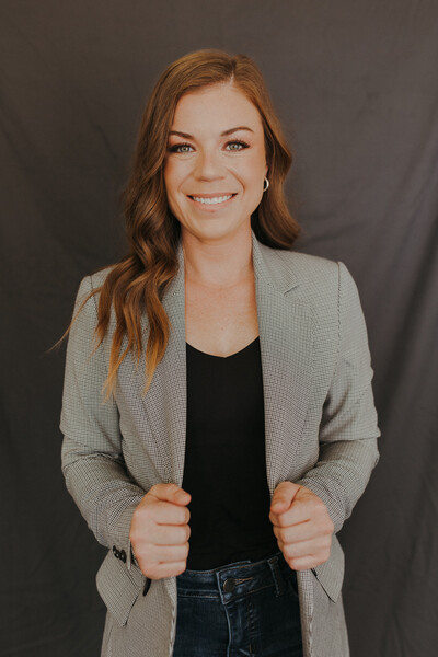 Team member of Nova Physiotherapy in Warman centered smiling. She is wearing a grey blazer, black shirt and jeans in front of black background.