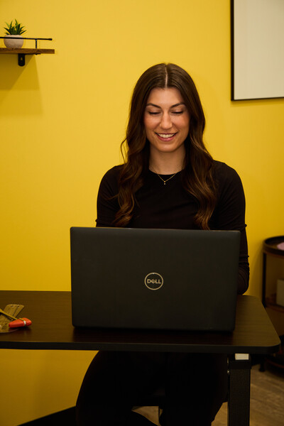 Team member of Nova Physiotherapy in Warman sitting at a desk in the clinic working on a laptop. She is smiling and demonstrating how to book online consultations.