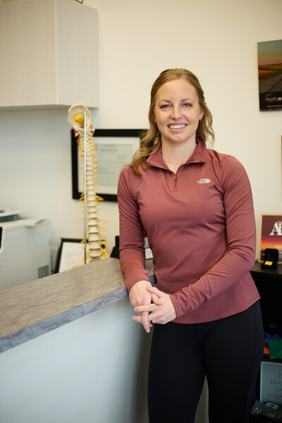Nova team member and physiotherpaist standing at front reception counter in clinic wearing pink 1/4 zip and model of spine in background