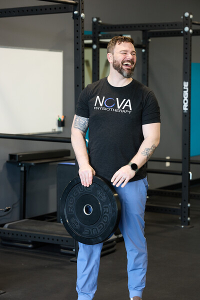 Team member and physiotherapist holding barbell weight plate and standing in gym with grey wall behind