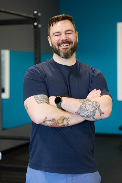 Nova team member and physiotherapist standing in clinic gym wearing blue shirt with blue wall and exercise equipment