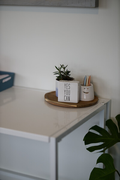 sign that reads yes you can on wooden plate with coloured pens and small plan ontop of white desk with plant beside