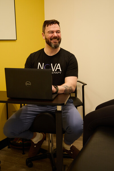 Team member and physiotherapist sitting on stool with laptop on desk wearing black nova shirt talking to person on treatment table