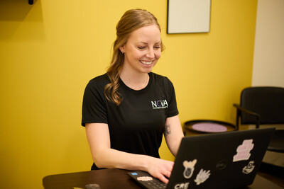 Woman wearing black nova shirt sitting at desk with computer and yellow wall behind her
